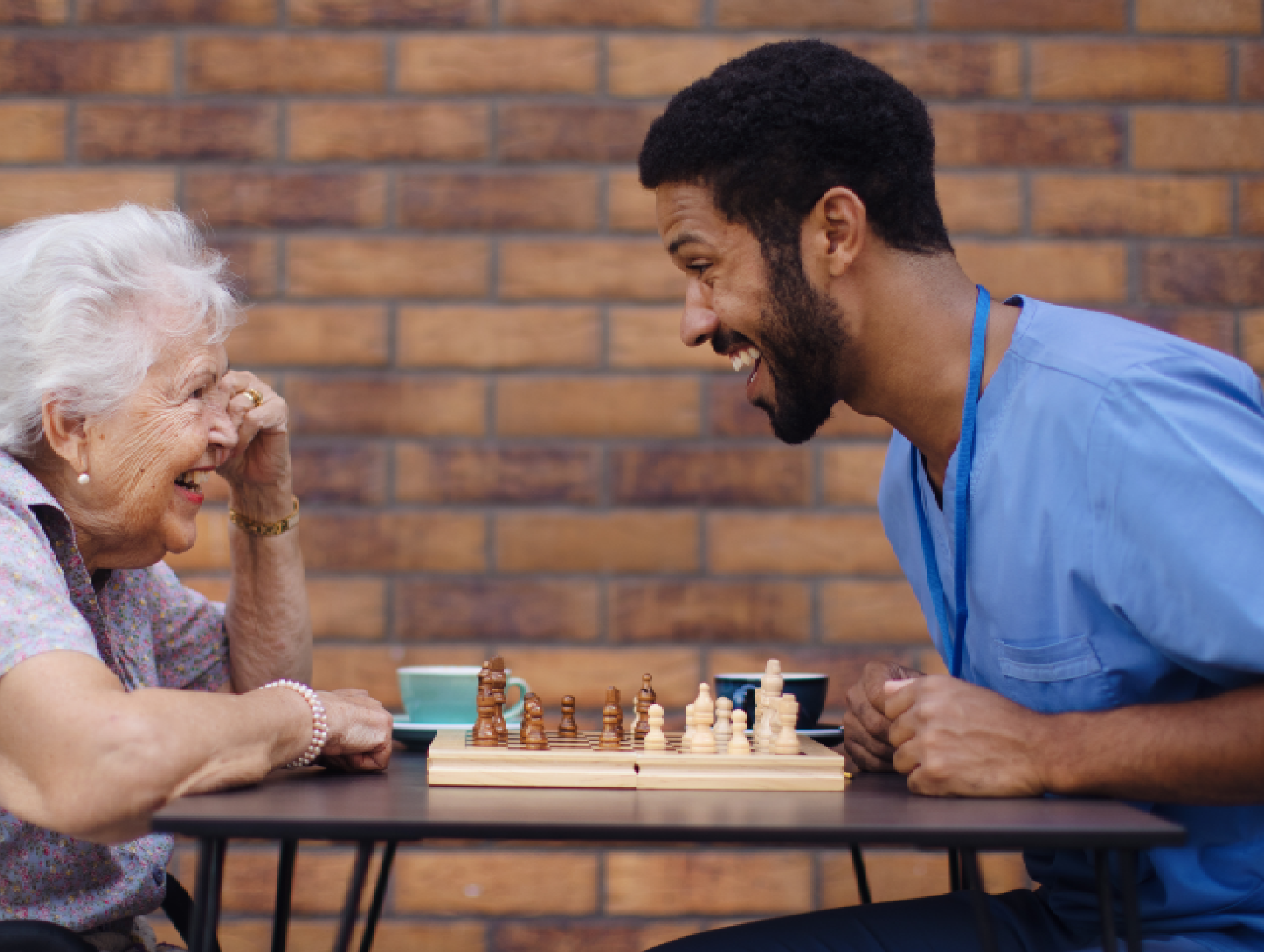 A professional caregiver is assisting a senior man with a simple puzzle, both smiling and engaged in a calm activity