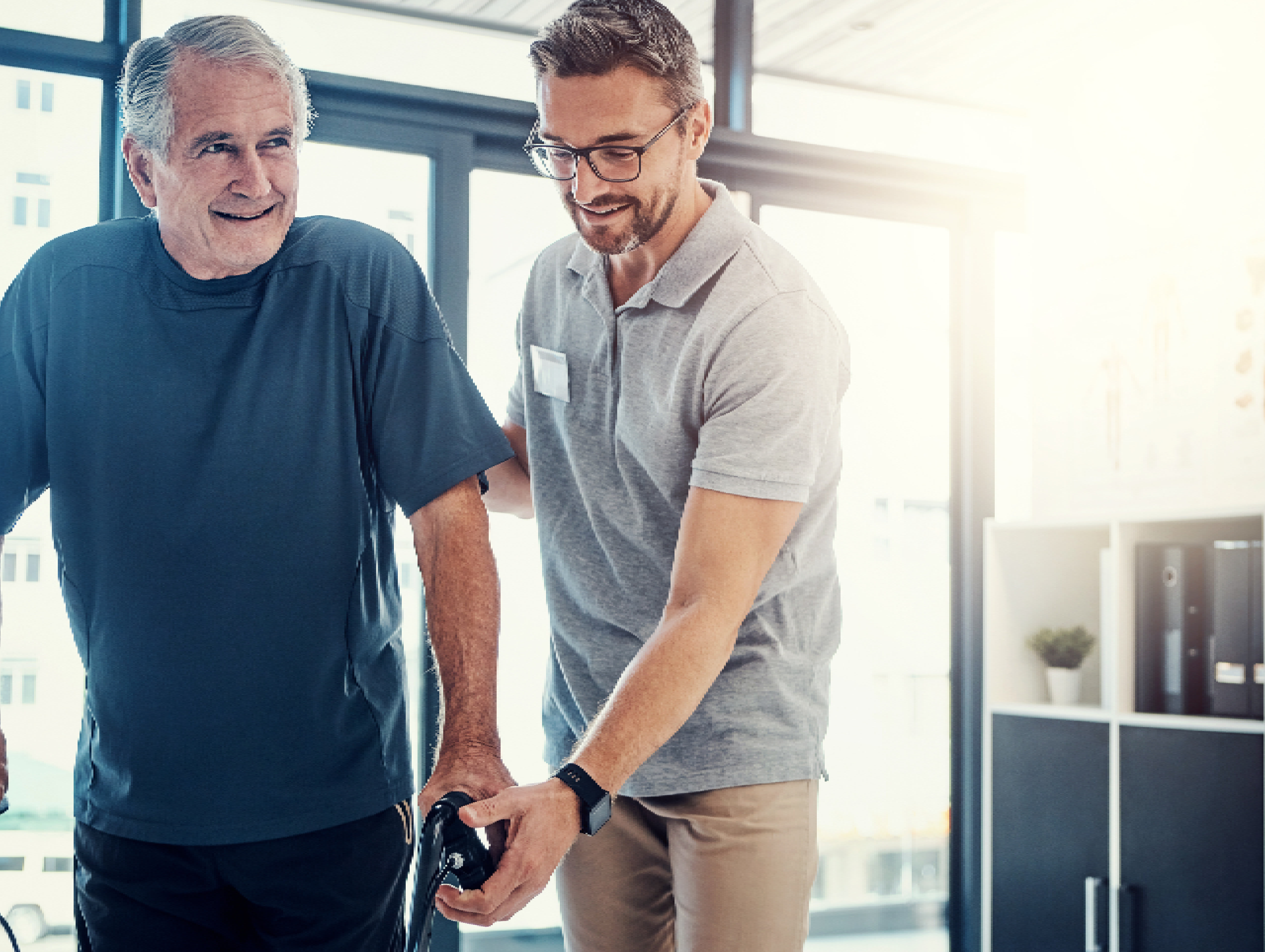 A professional physiotherapist is helping a senior man to walk with a frame in a clean, bright hallway, showing mobility assistance.