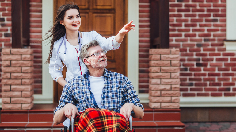 A professional caregiver assisting a senior woman with gentle mobility in a brightly lit hallway of an Assisted Living home for elderly in India